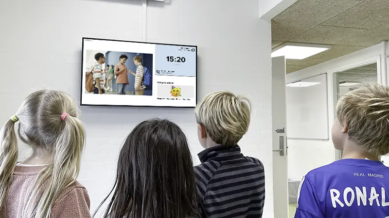 Photo of a digital signage screen. A group of students is looking at the content, which displays a calendar post, a clock, and a birthday greeting.