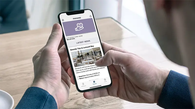A person holding a mobile phone and viewing news and updates in the PinToMind Go app, specifically for relatives at a care home. Photo.