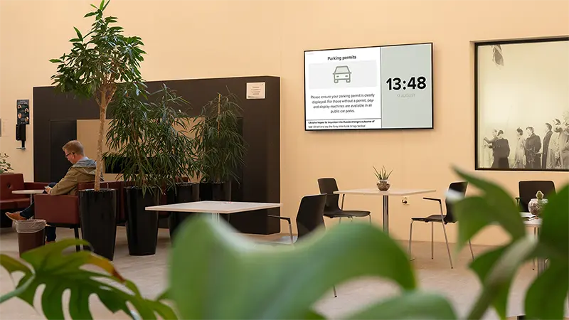 A large information screen on the wall in a bright and welcoming hospital waiting area displays the time and a post about parking permits. A man is seated in the lounge area. Photo.