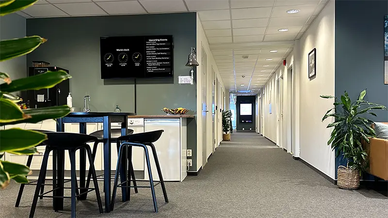 An information screen displaying a world clock post and upcoming events on the wall in an office space, with seating and a kitchen counter in the foreground.