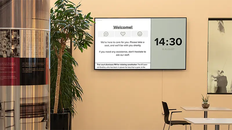 A screen in a modern waiting area displays a welcome message, current time, and a news ticker. Photo