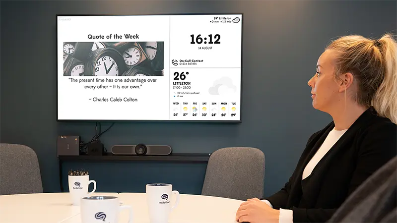 A woman sits at a table in the teachers' lounge, looking up at a screen displaying the Quote of the Week, time, and weather forecasts. Photo.