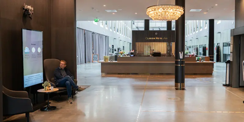 A man is sitting in a hotel lobby, looking at an information screen that displays weather forecasts. Photo.